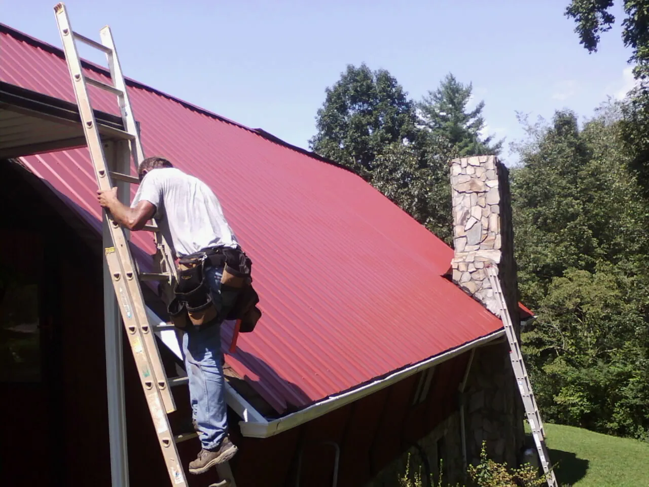 Roofer on a ladder beside a red metal roof and stone chimney — Soffit & Fascia Repair by Roofing Brothers in Brownsboro